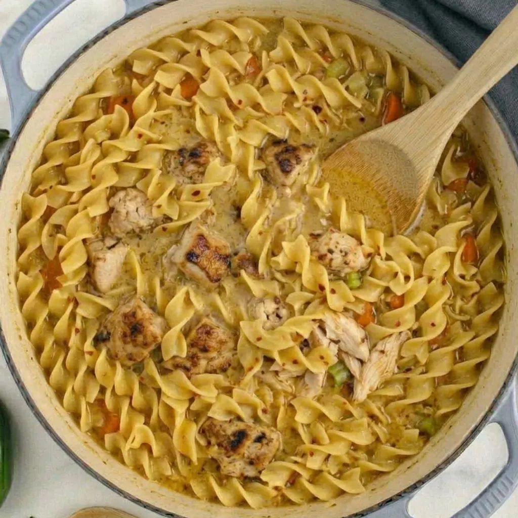 Spicy chicken noodle soup in a large Dutch oven, packed with rotini pasta, seared chicken chunks, red pepper flakes, and vegetables, with a wooden spoon dipping in.