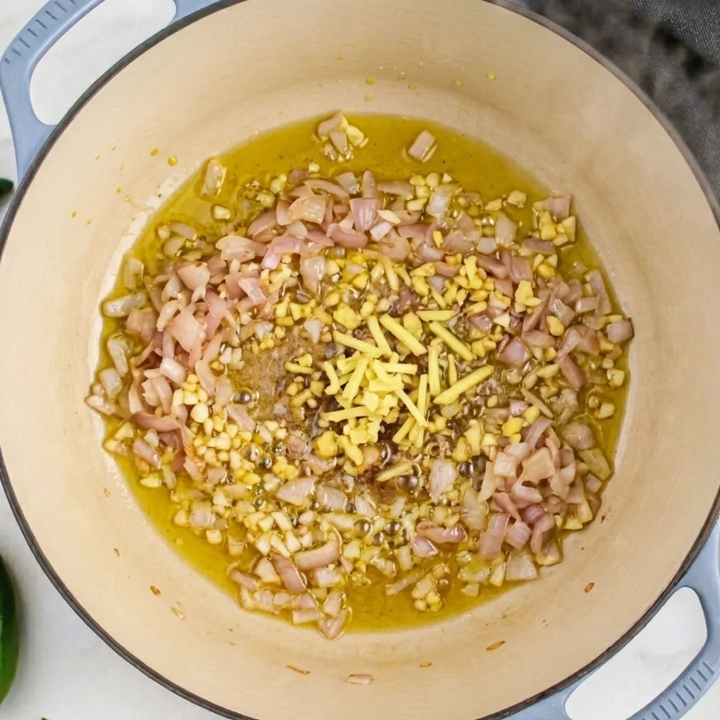 Sautéing aromatics for spicy chicken noodle soup, featuring diced onions, minced garlic, and fresh ginger matchsticks sizzling in olive oil inside a blue Dutch oven.