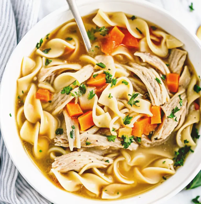 A white bowl filled with spicy chicken noodle soup, featuring shredded chicken, wide egg noodles, diced carrots, and chopped parsley in a flavorful, golden broth with a spoon.