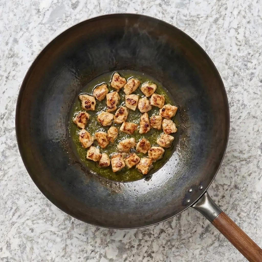 seasoned cubes of chicken breast are searing in hot oil at the center of a dark wok, set against a light marbled countertop."