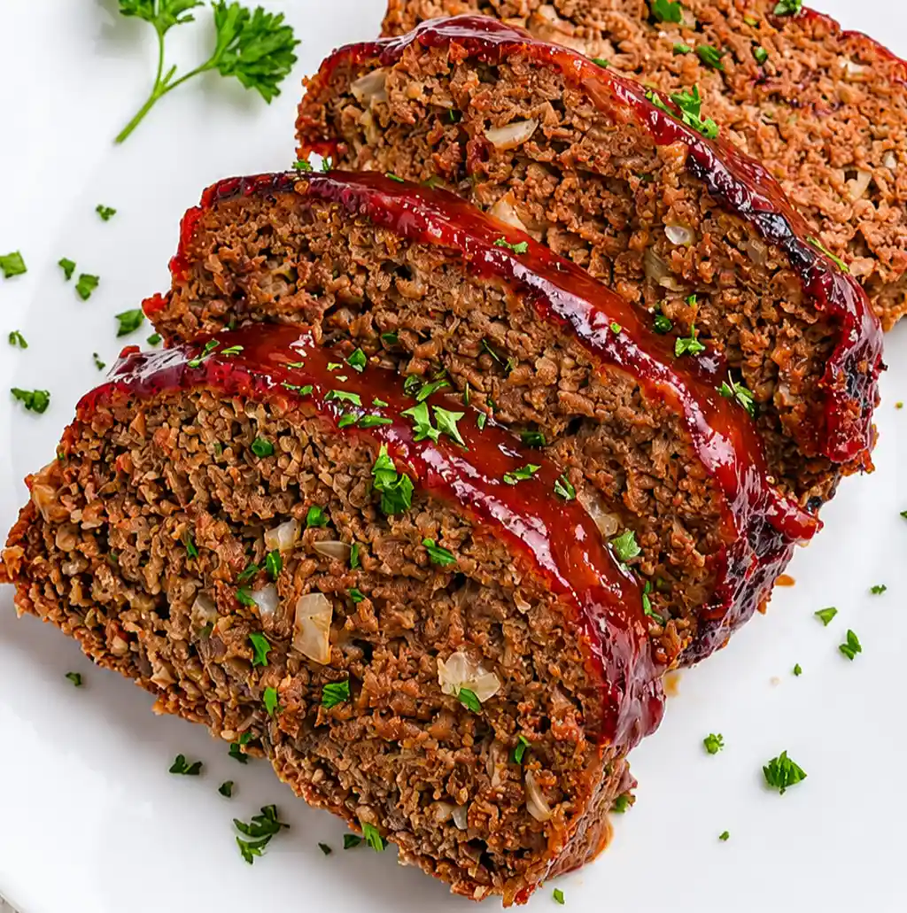 Slices of juicy, cooked Air Fryer Meatloaf Recipe featuring a glossy, caramelized red glaze and visible onion bits, served on a white platter garnished with fresh chopped parsley.