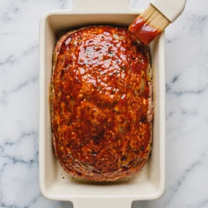 A pastry brush is used to spread a thick layer of red sauce on French Onion Meatloaf in a casserole dish, which is placed on a marble countertop.