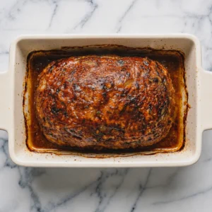 Overhead shot of a fully cooked, glossy glazed French onion meatloaf baked to a golden-brown finish inside a cream-colored ceramic baking dish on a marble countertop.